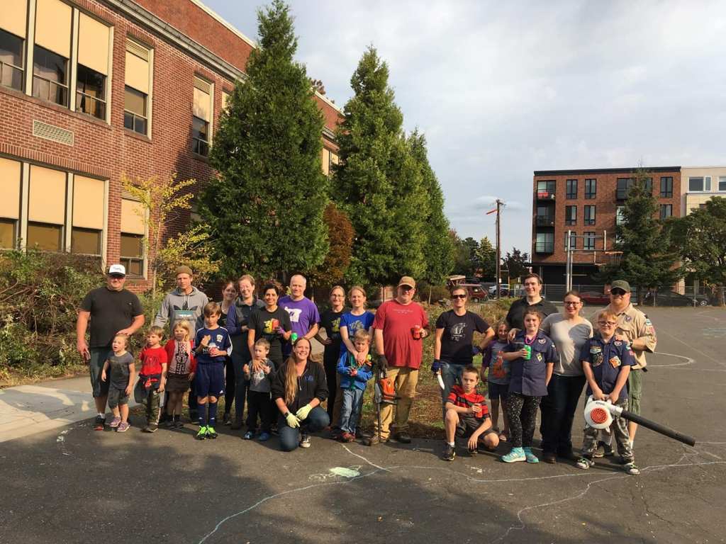 A photo of a group of families standing and sitting in the James John playground. Some have gardening clothes on and one child is holding a leaf blower.